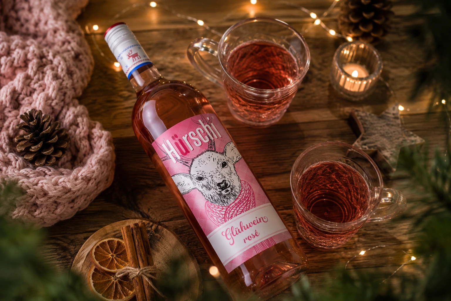 Hürschi Glühwein Rosé liegend auf einem weihnachtlich gedeckten Holztisch mit Glühweinbechern, Lichterkette, Zimt, Orangenscheiben und winterlicher Dekoration.