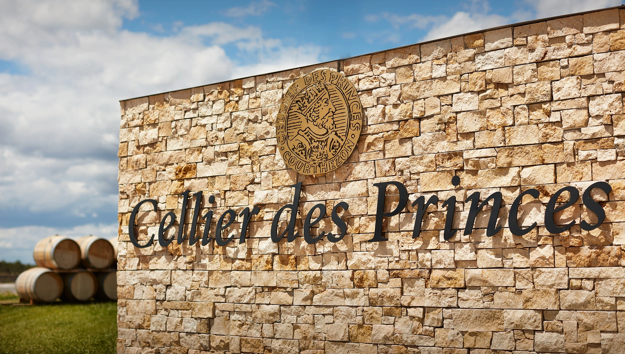 Eingangsbereich von Cellier des Princes mit Schriftzug auf Steinmauer, blauem Himmel und Weinfässern im Hintergrund