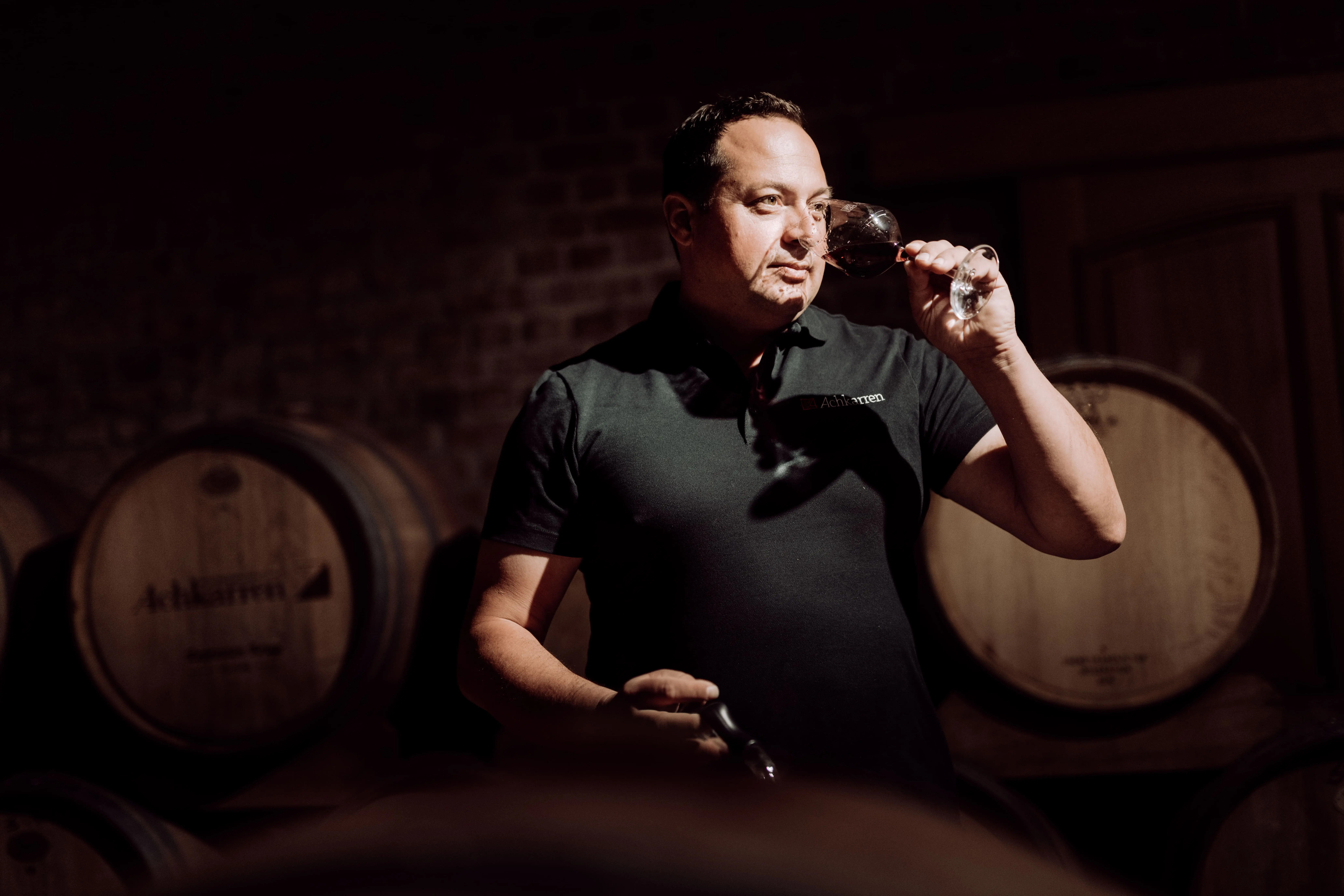 Winzer der Winzergenossenschaft Achkarren riecht an einem Glas Wein, im Hintergrund Achkarren Weinfässer