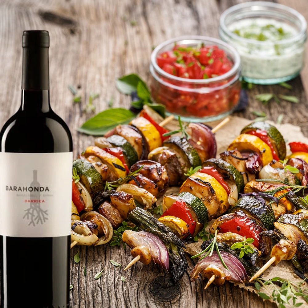 Flasche Barahonda Barrica Rotwein auf einem rustikalen Holztisch, serviert mit gegrilltem Fleisch und verschiedenen Dips in kleinen Schalen, warme gesellige Atmosphäre im Outdoor-Bereich.