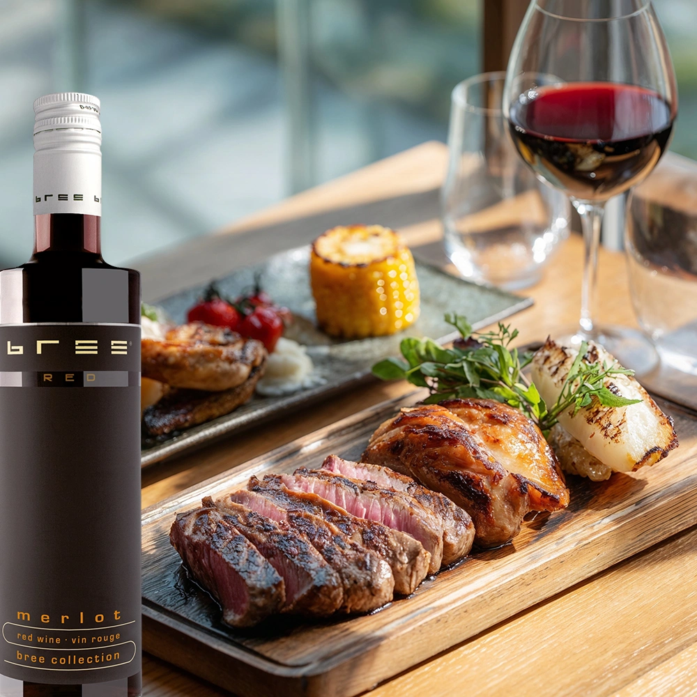 Kleine 0,25-Liter-Flasche Bree Merlot auf einem gedeckten Tisch mit gefülltem Rotweinglas, geschnittenen Steakstreifen und aromatischem Grillgemüse als Speisebegleitung