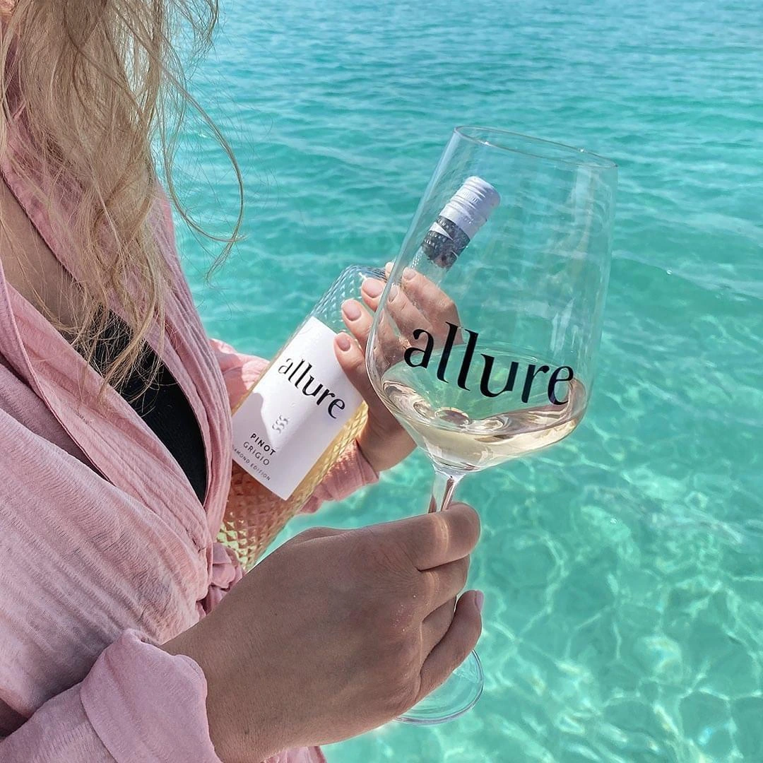 Frau hält eine Flasche Allure und ein Glas Wein, im Hintergrund türkisblaues Meer
