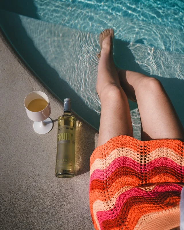 Flasche Bree Weißwein am Pool neben einer Frau mit Füßen im Wasser, sommerliche und entspannte Urlaubsatmosphäre
