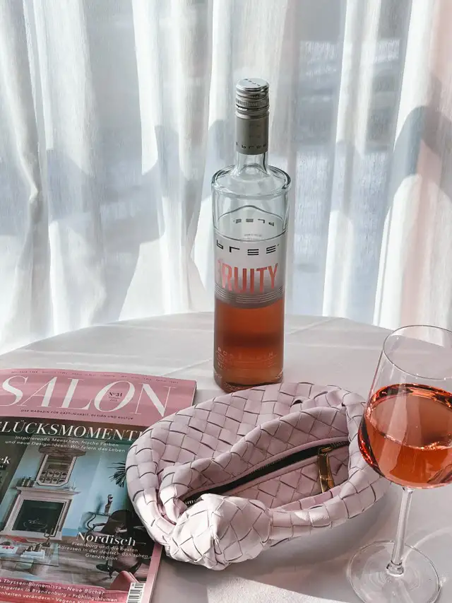 Bree Fruity Rosé Wein auf einem Tisch mit gefülltem Roséglas, Handtasche und Zeitschrift neben einer lichtdurchfluteten Gardine in sommerlicher Atmosphäre