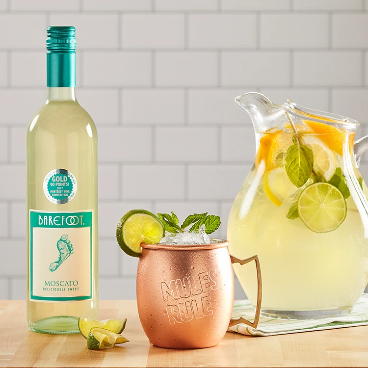 Barefoot Moscato Flasche auf Tisch mit Karaffe voller Zitrusfrüchte und Bronzebecher im Vordergrund